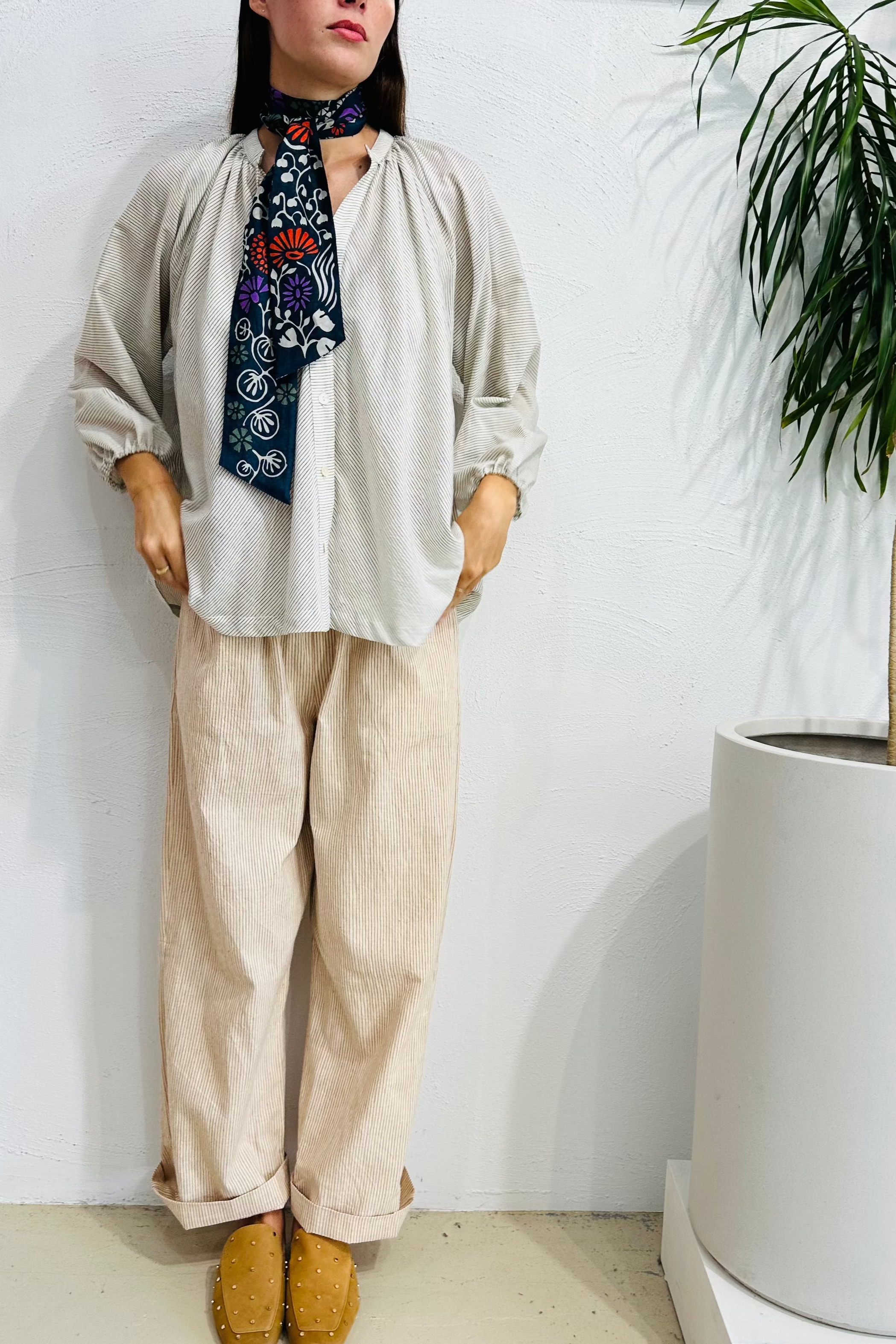 Person wearing a beige outfit with a patterned scarf, standing indoors with a plant in the background.