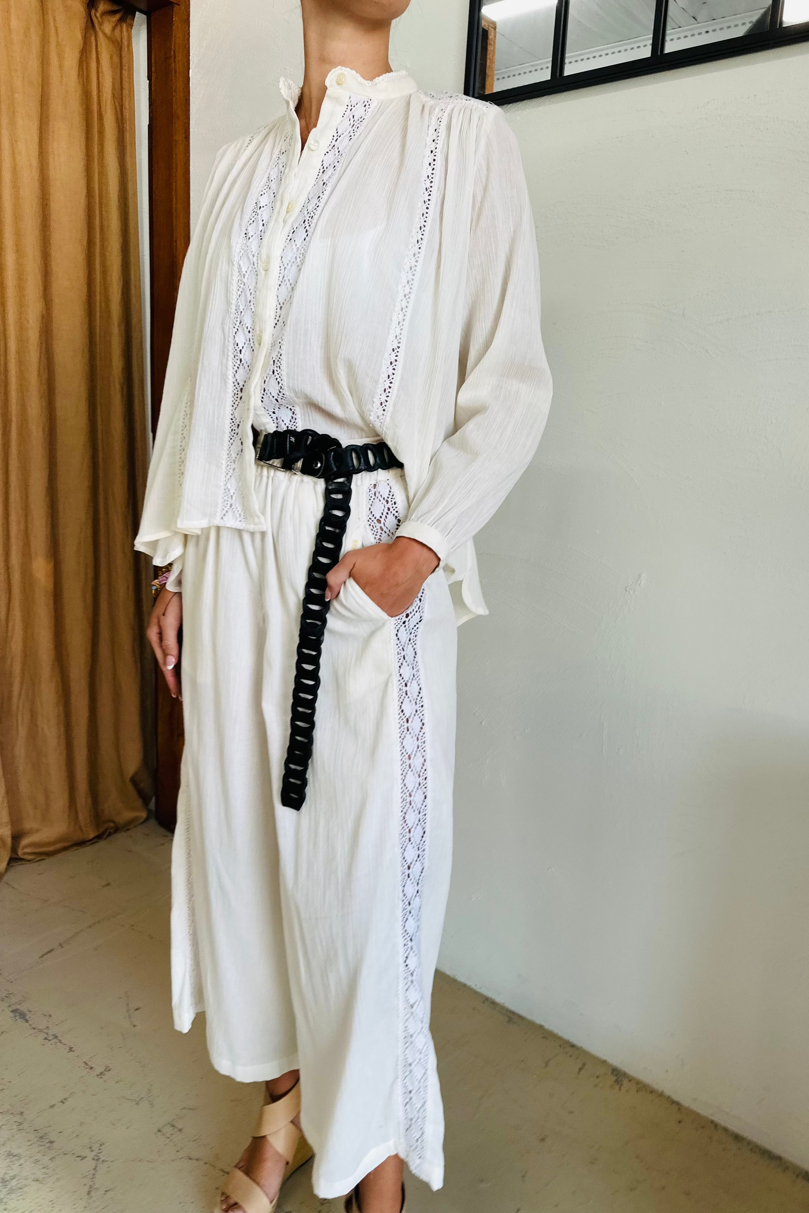 Person wearing a white outfit with lace details and a black belt in a minimal indoor setting.